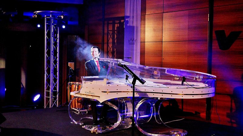 Alex Parker sitzt im Scheinwerferlicht an einem transparenten Flügel und singt